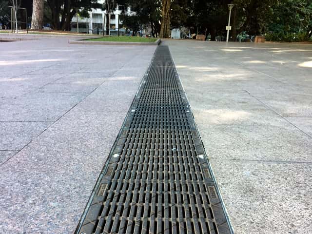 Long trench drain grate installed across a large stone tiled commercial plaza Long trench drain grate installed across a large stone tiled commercial plaza