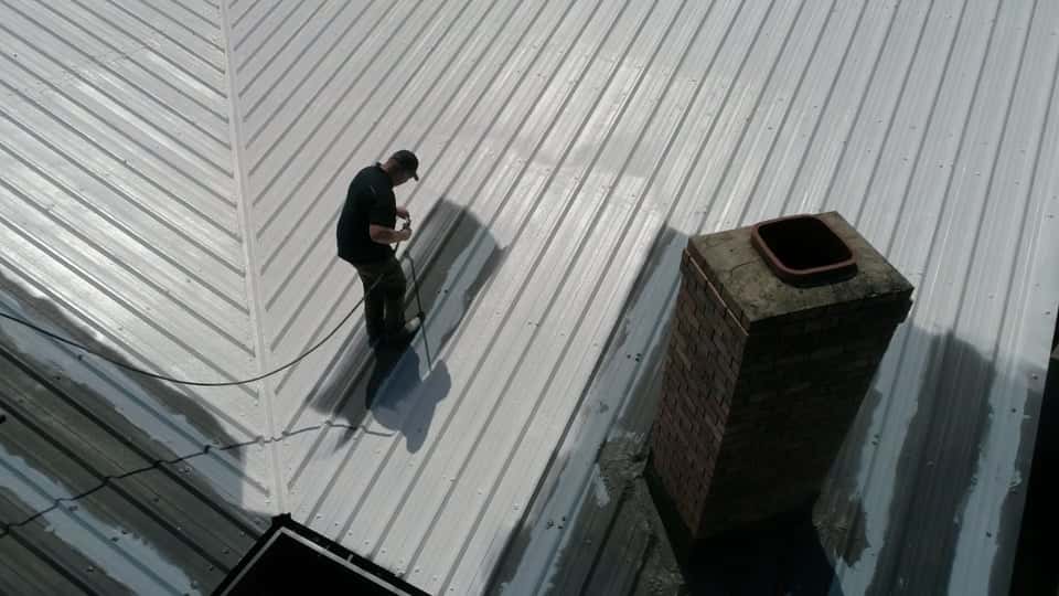 Roofer spray-applying a reflective white elastomeric coating on a corrugated metal roof near a brick chimney Roofer spray-applying a reflective white elastomeric coating on a corrugated metal roof near a brick chimney