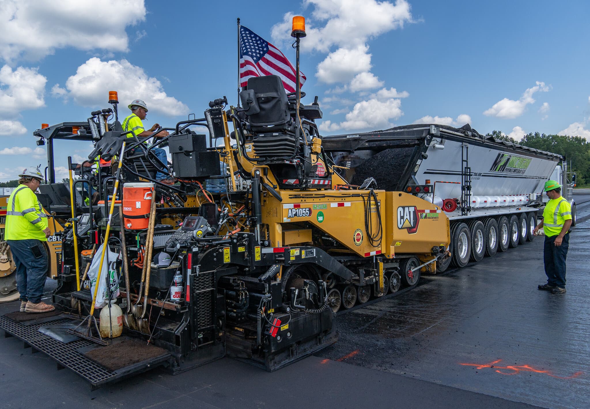 Patriot Asphalt & Construction — Albuquerque, New Mexico Road construction crew operating a yellow CAT asphalt paver machine with an American flag attached, paving a highway under a blue sky