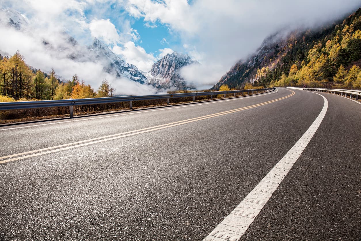 Winding two-lane asphalt highway curving through misty snow-capped mountains surrounded by autumn forest foliage Winding two-lane asphalt highway curving through misty snow-capped mountains surrounded by autumn forest foliage