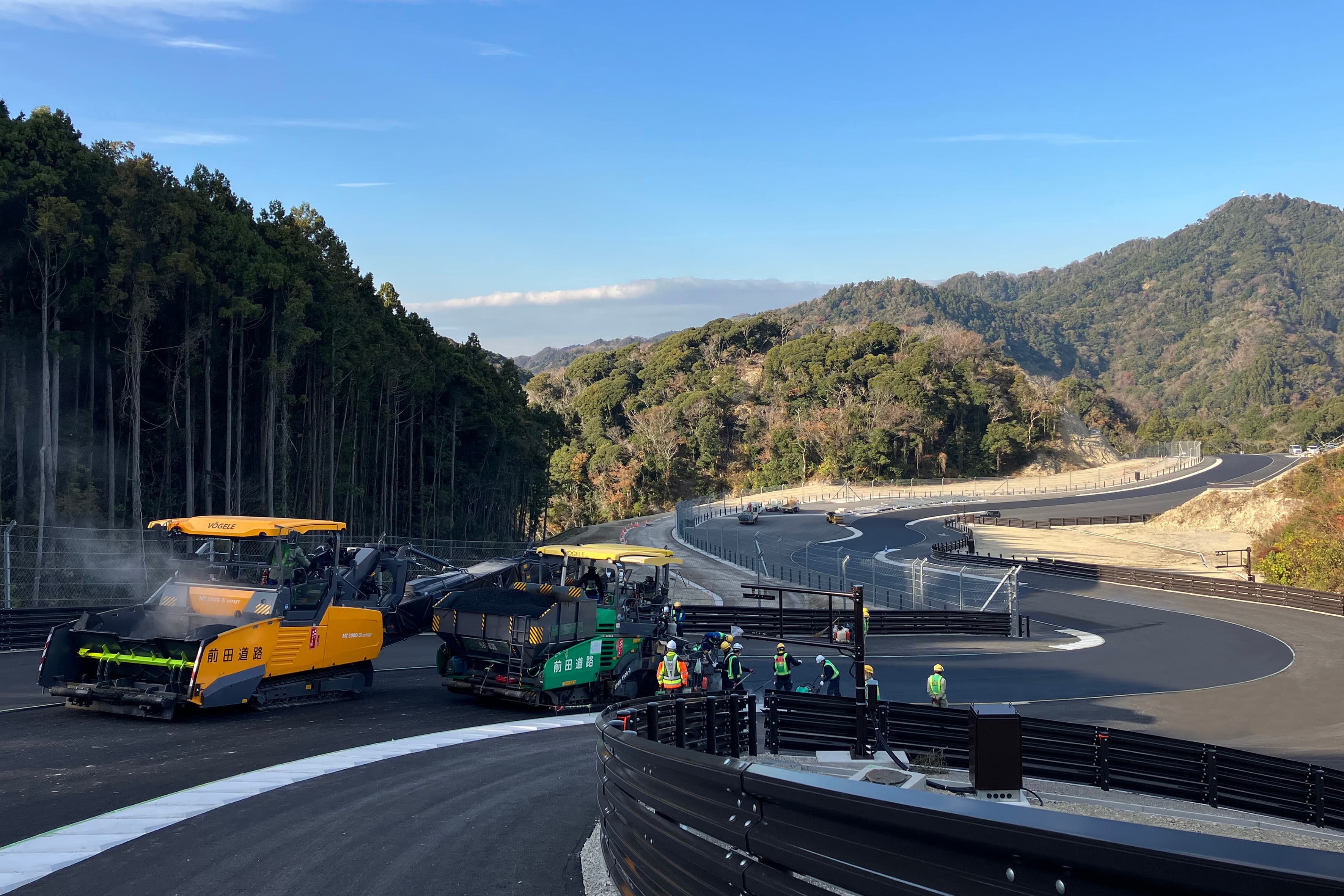 Asphalt Paving and Rollers Heavy asphalt paving machinery laying fresh blacktop on a winding curved road along a forested mountain slope